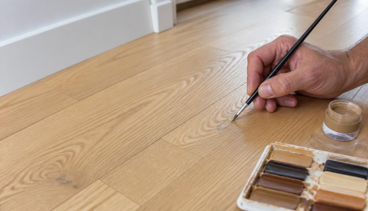 Technician colour-matching a localised repair on a timber-effect hard floor using a fine brush and tinted finishes.