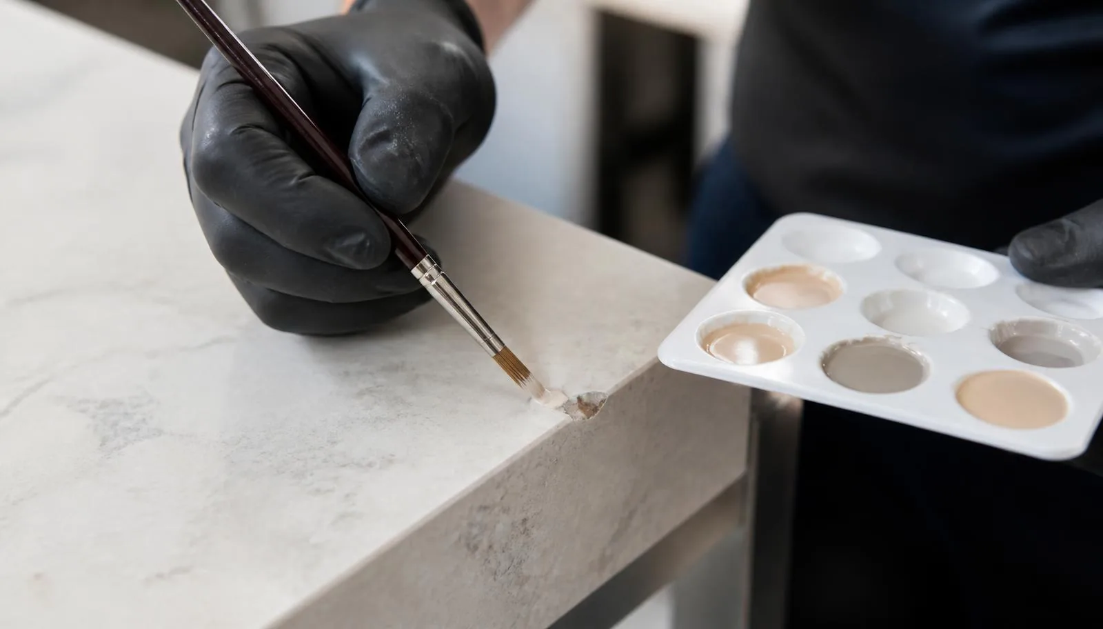 Close-up of a technician brushing colour-matched filler into a chipped stone-effect worktop edge, with a palette of tinted resins alongside.